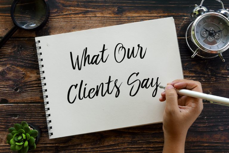 a hand holding a pen and writing "what our clients say" on the note pad situated at the table