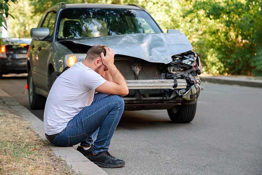 A man sitting on a road gutter in front of a wrecked car after a car accident.